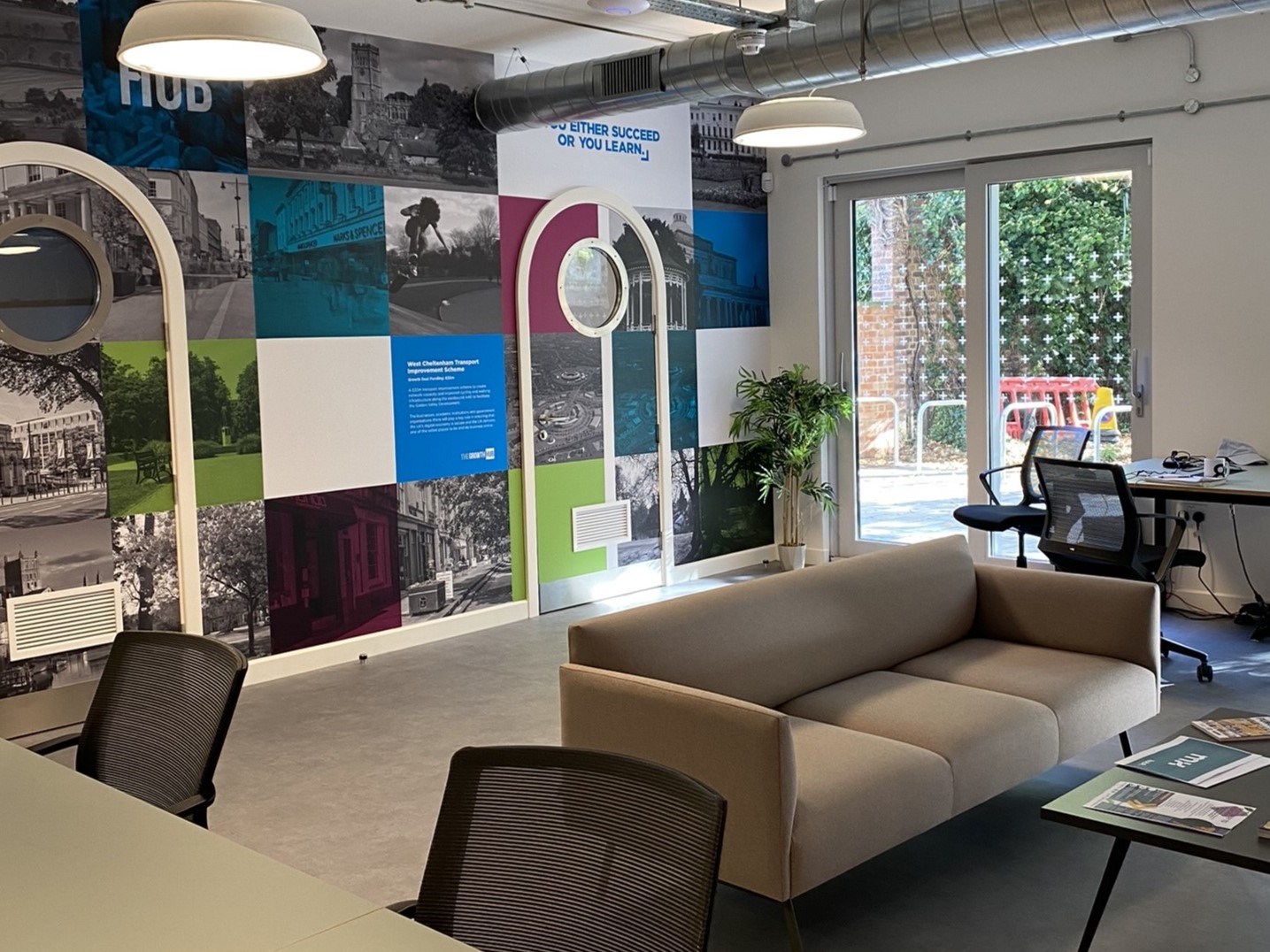 The image shows a co-working space with a colourful chequerboard of imagery across the back wall, with 2 doors leading to small meeting rooms. A sofa, coffee table, desks and chairs are in the foreground.