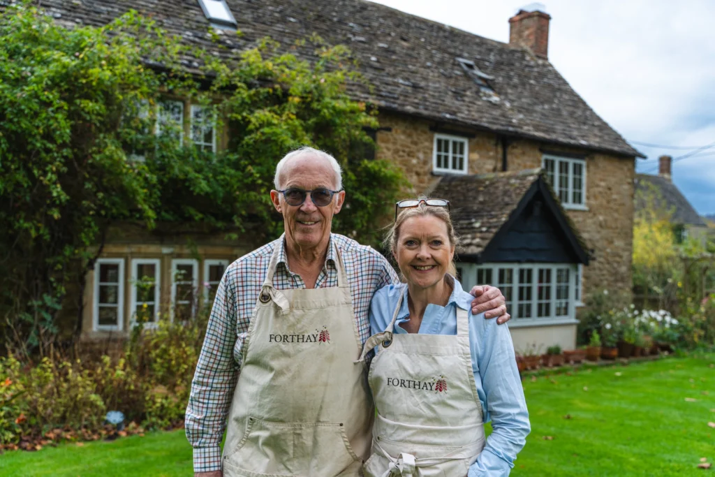 The image shows a country cottage in the background with its owners, husband and wife, Debs and Charles Lamplugh in the foreground smiling at the camera.