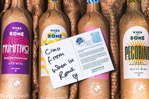 Postcard saying ciao from When in Rome on top of bottles of wine